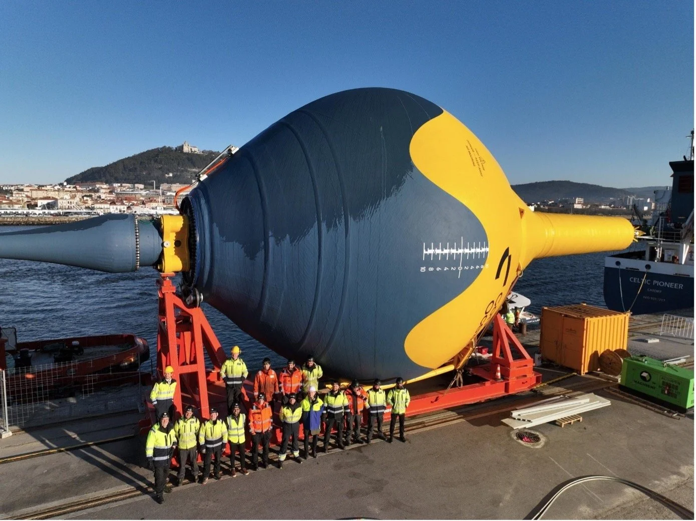 A large submarine on a shipping dock with workers standing in front, with a cityscape and hills in the background.