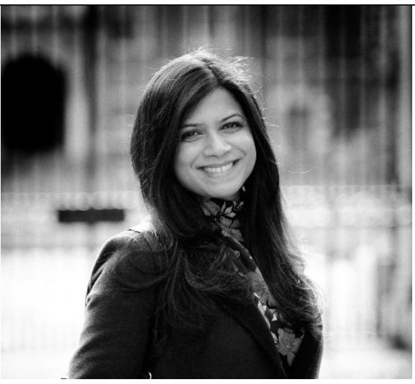 Black and white photo of a woman with long dark hair, smiling, wearing a blazer and floral top, outdoors with blurred background.