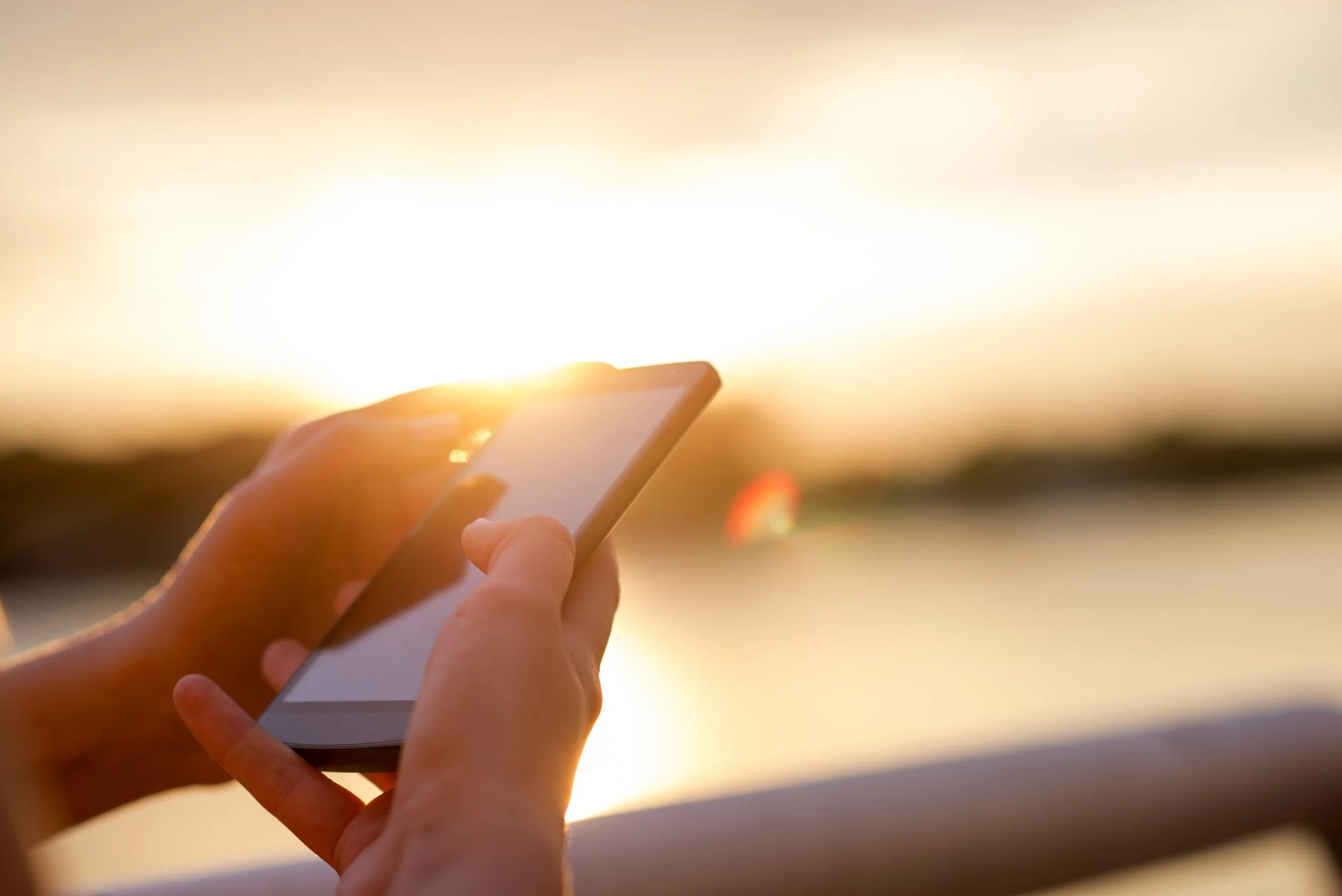 Person holding a smartphone outdoors at sunset, with the sun shining brightly in the background