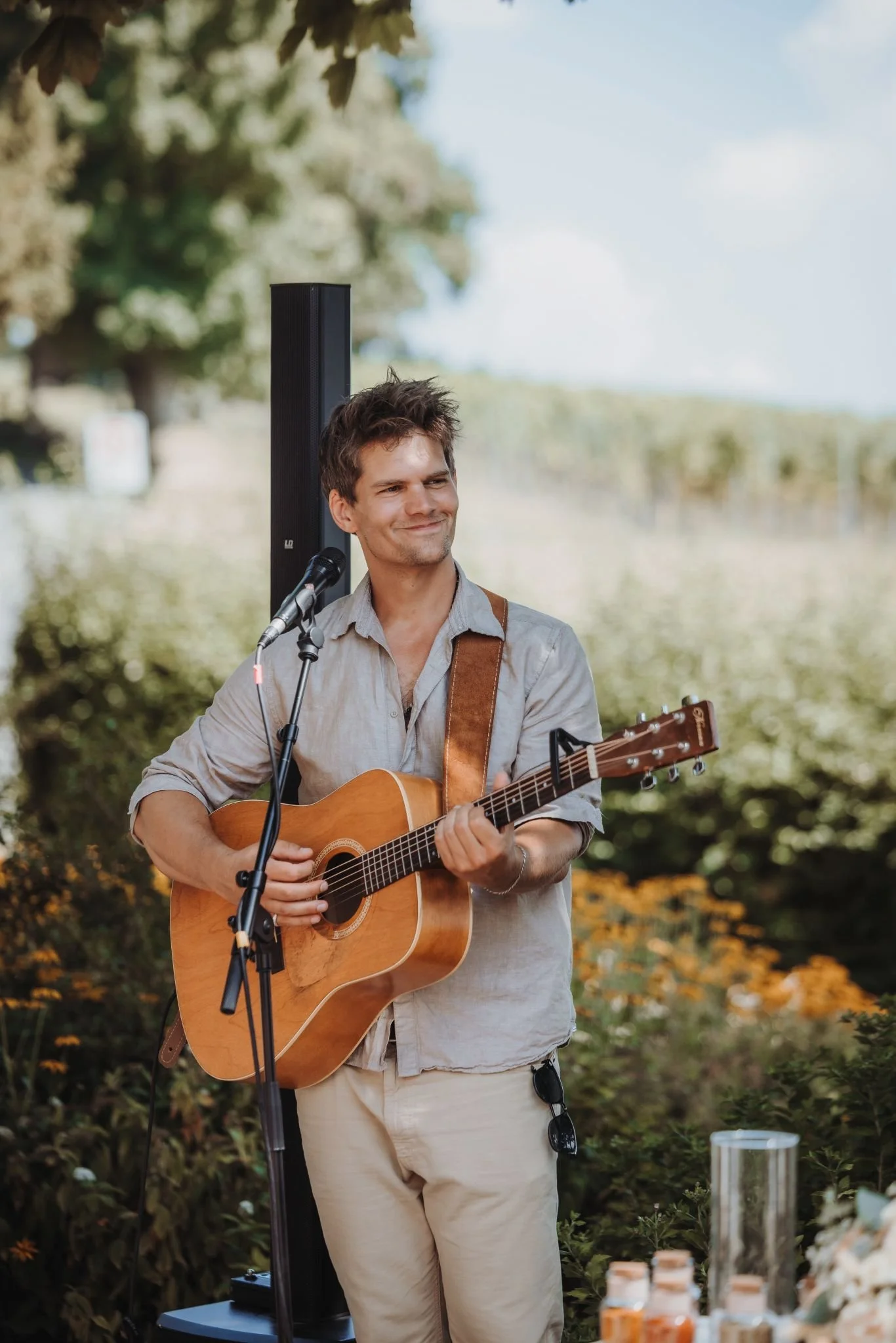 A man playing an acoustic guitar outdoors with a microphone in front of him, surrounded by nature and flowering plants.