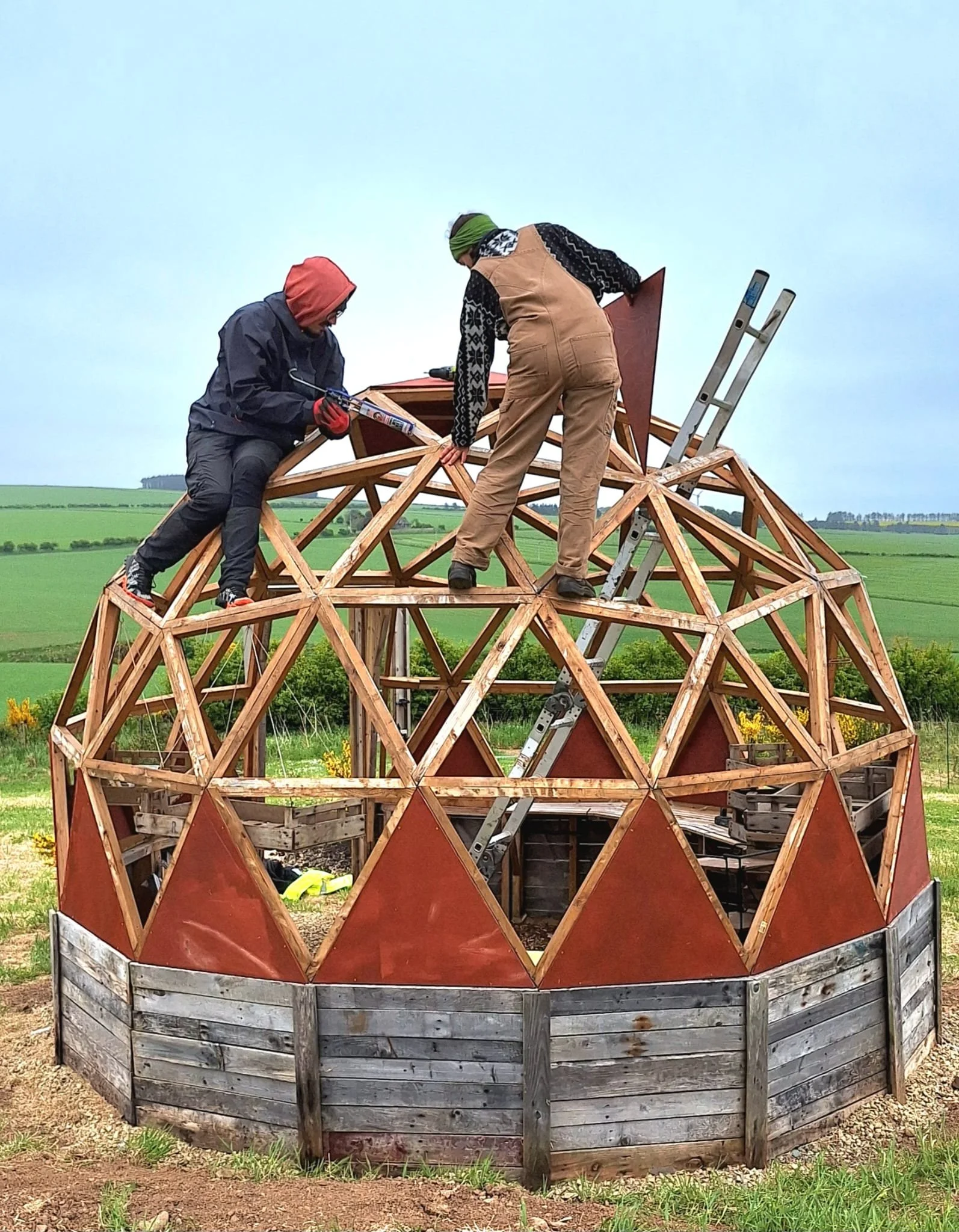 Two people working on the construction of a geodesic dome structure outdoors with green fields in the background.