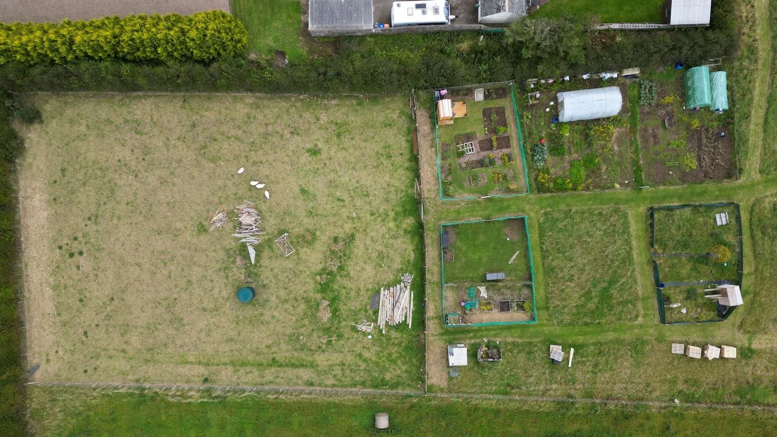 Aerial view of a backyard with multiple garden plots, a large patch of grass with some debris, and fenced areas with garden beds and small structures.
