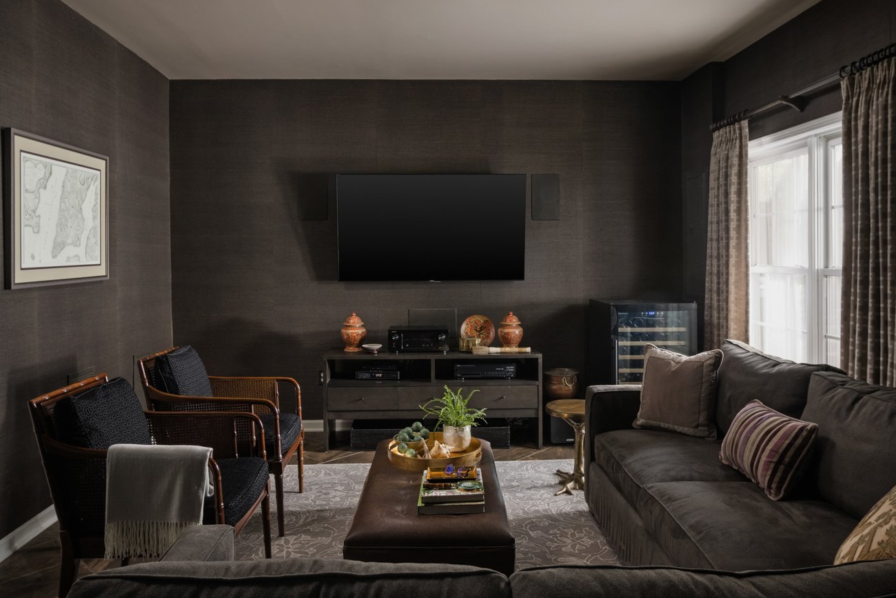 Living room with dark walls, a mounted flat-screen TV, a gray sofa with patterned pillows, two armchairs, a small table with decor, a wine cooler, and a window with curtains.