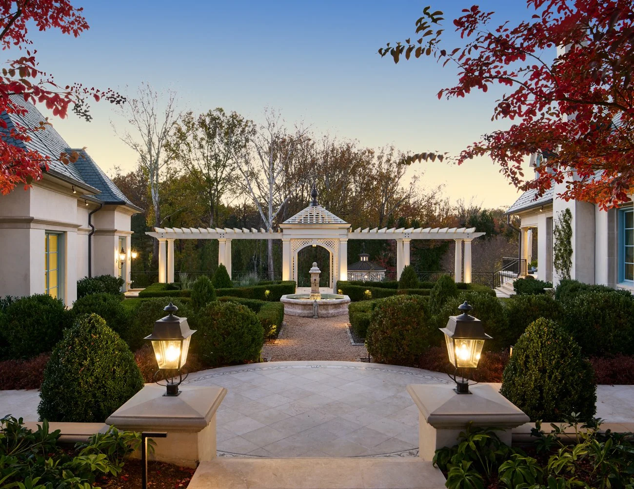 Elegant courtyard garden with fountain, surrounded by manicured hedges and classical architectural structures, lit by lanterns at dusk.