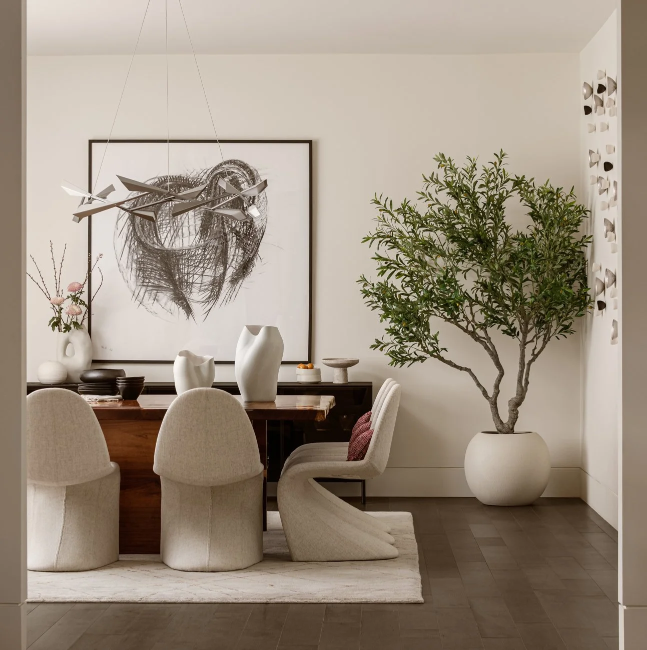 A dining room with a large leafy potted plant, abstract art on the wall, cream upholstered chairs, and a wooden dining table decorated with vases and bowls.