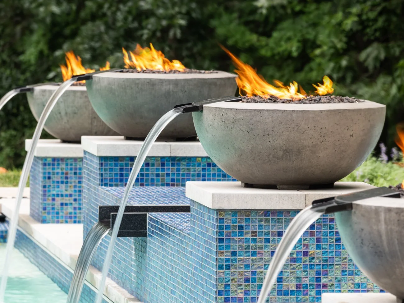 Outdoor water fountain with concrete bowls emitting flames, set against greenery. The fountain is decorated with blue mosaic tiles.
