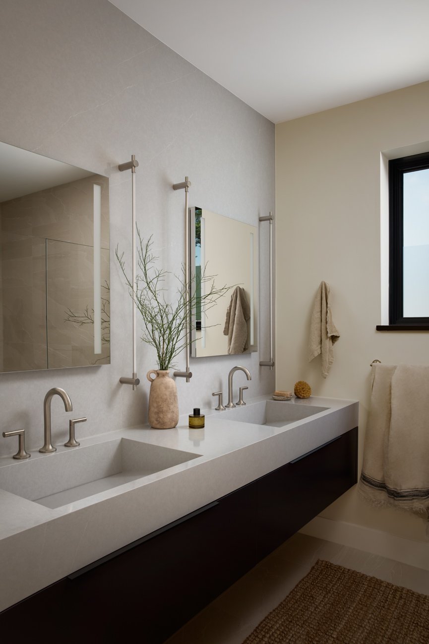 Modern bathroom with dual sinks, large mirror, beige towels, glass vase with plant, window, and minimal decor.