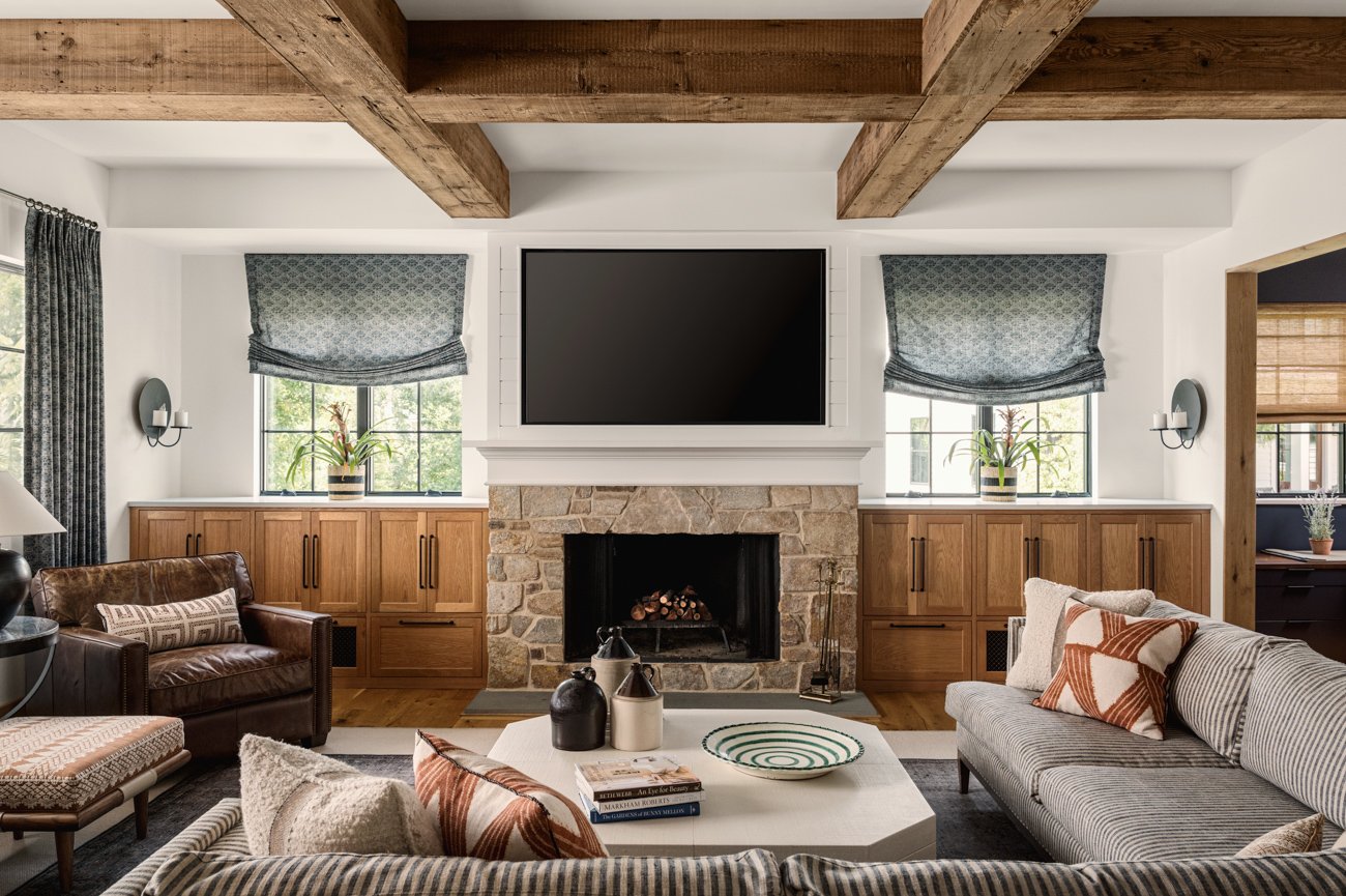 Living room with a stone fireplace, flat-screen TV above, wooden beam ceiling, two windows with blue patterned curtains, and a sectional sofa with decorative pillows.