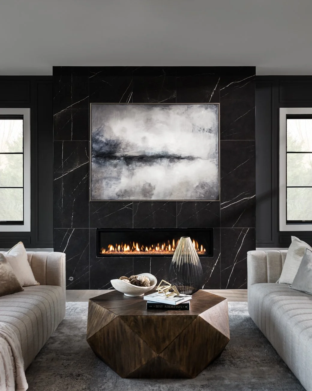 Living room with a modern black marble fireplace, abstract black and white painting above, beige sofas, a wooden coffee table, and decorative items, with two windows on either side.