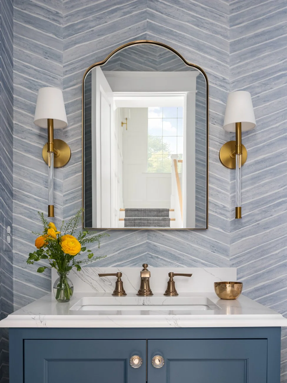 A bathroom vanity with a blue cabinet, a white marble countertop, and a rectangular mirror. There are two wall-mounted brass sconces, a vase with yellow flowers, and a small brass bowl.