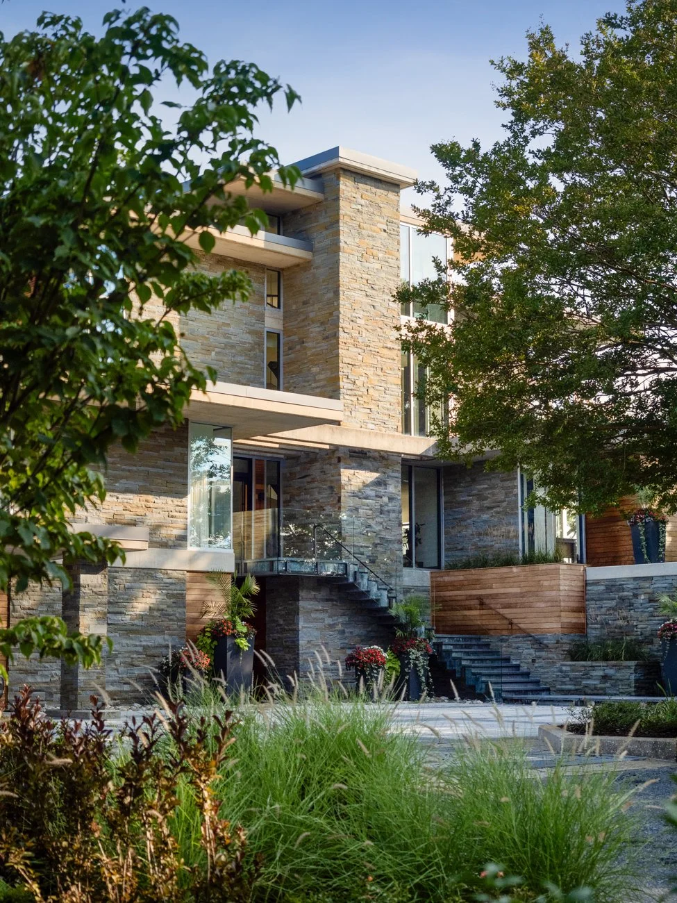Modern multi-story house with a stone exterior, large glass windows, and surrounding greenery including trees and plants.