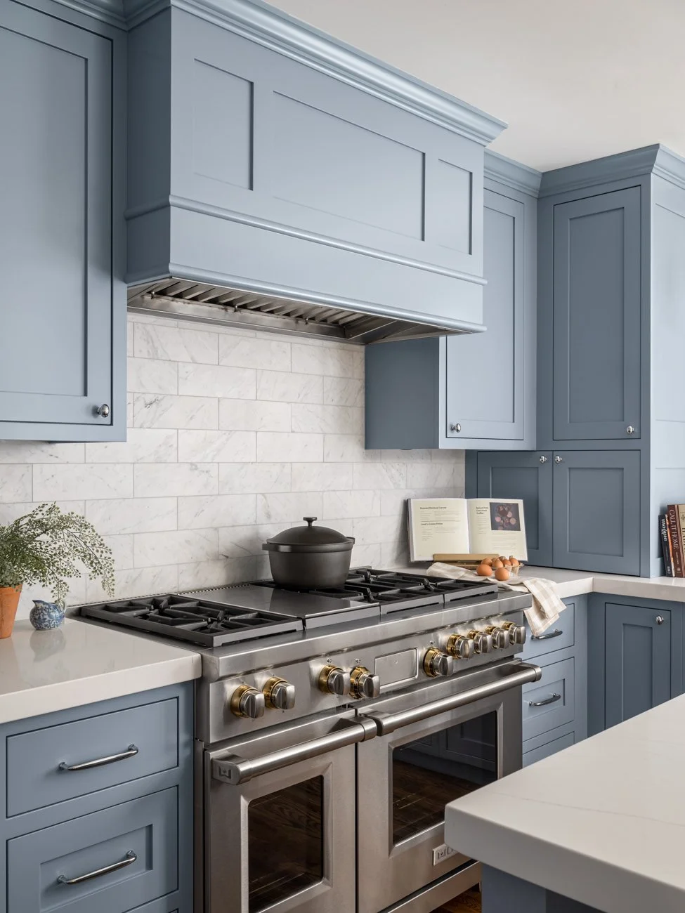 Kitchen with blue cabinets, white countertops, stainless steel stove, and a tiled backsplash. There are a pot on the stove, a book, eggs, and some decorative items on the counter.