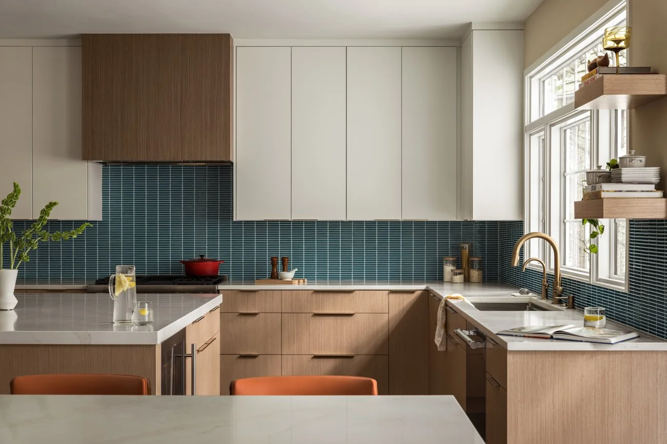 Modern kitchen with wood and white cabinetry, blue tiled backsplash, and a window in the background. On the counter, there are plants, a glass of water with lemon, and a red pot on the stove.