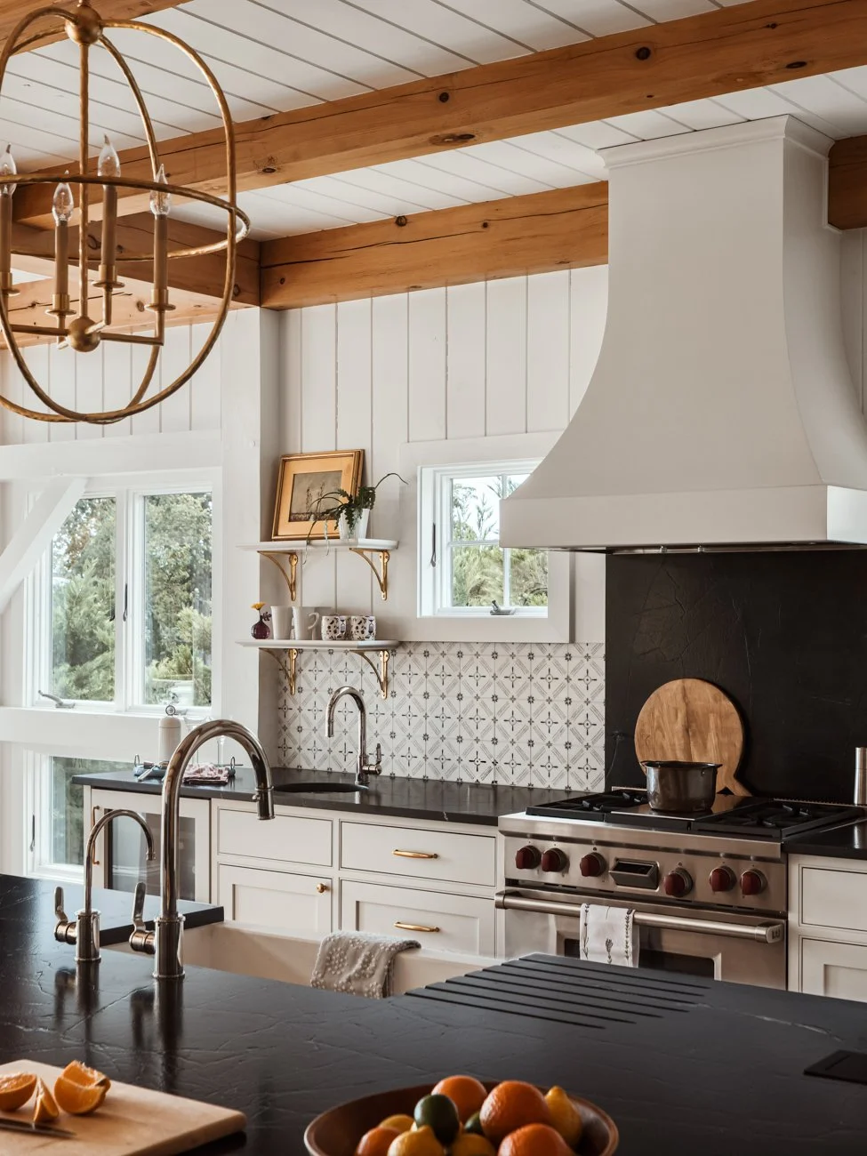A rustic kitchen with white cabinets, black countertops, a stainless steel stove, and a white range hood. Wooden beams and white shiplap walls with windows overlooking greenery.
