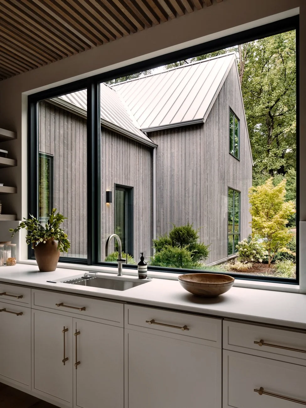 Kitchen window view showing a modern house with vertical wood siding, large windows, and a metal roof, surrounded by trees and greenery.