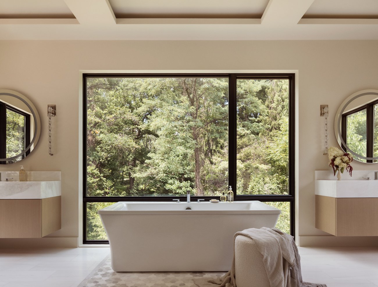 Modern bathroom with a bathtub in front of large window overlooking green trees, with two floating vanities with sinks and round mirrors on either side.
