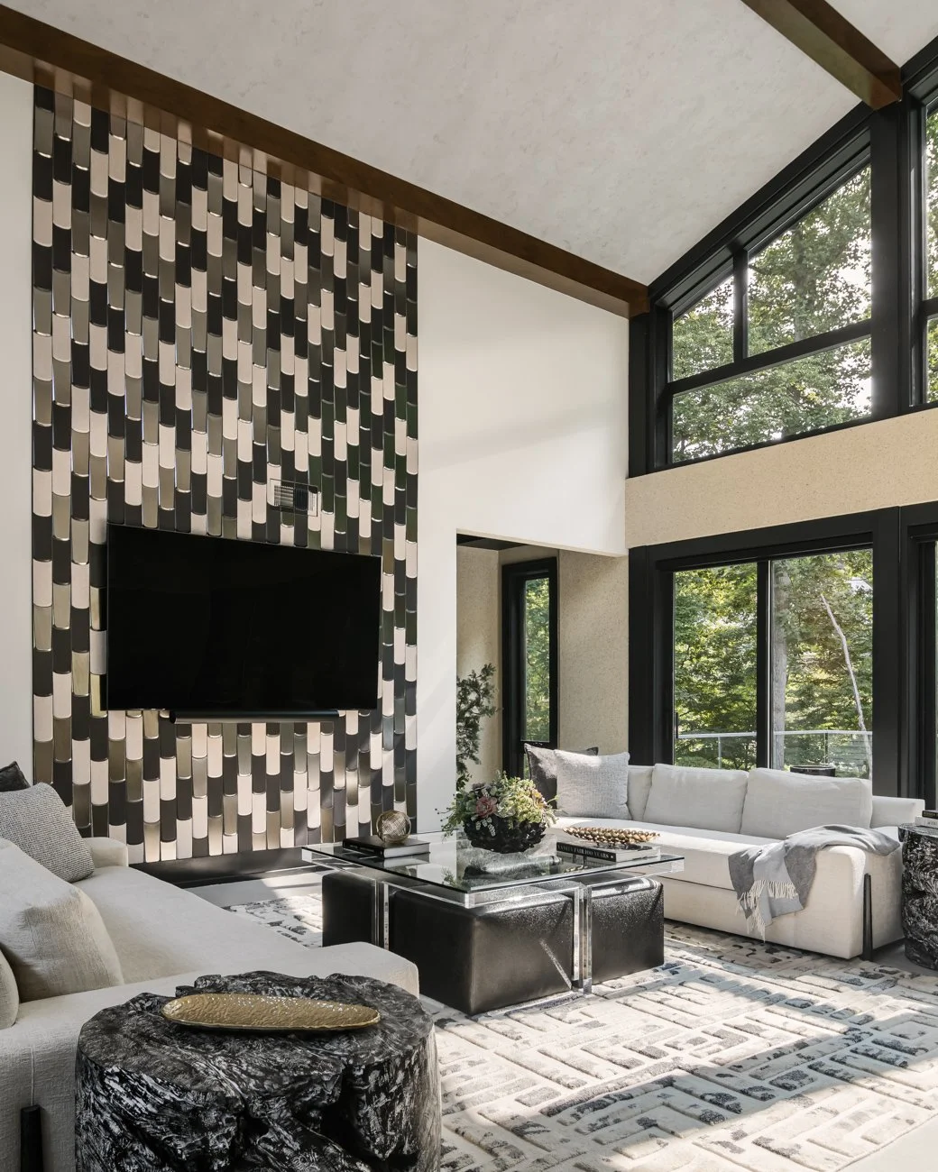 Contemporary living room with high ceiling, large windows, white sofa, black and white patterned carpet, modern coffee table, and a black accent wall with a display of vertically oriented rectangular panels and a wall-mounted TV.