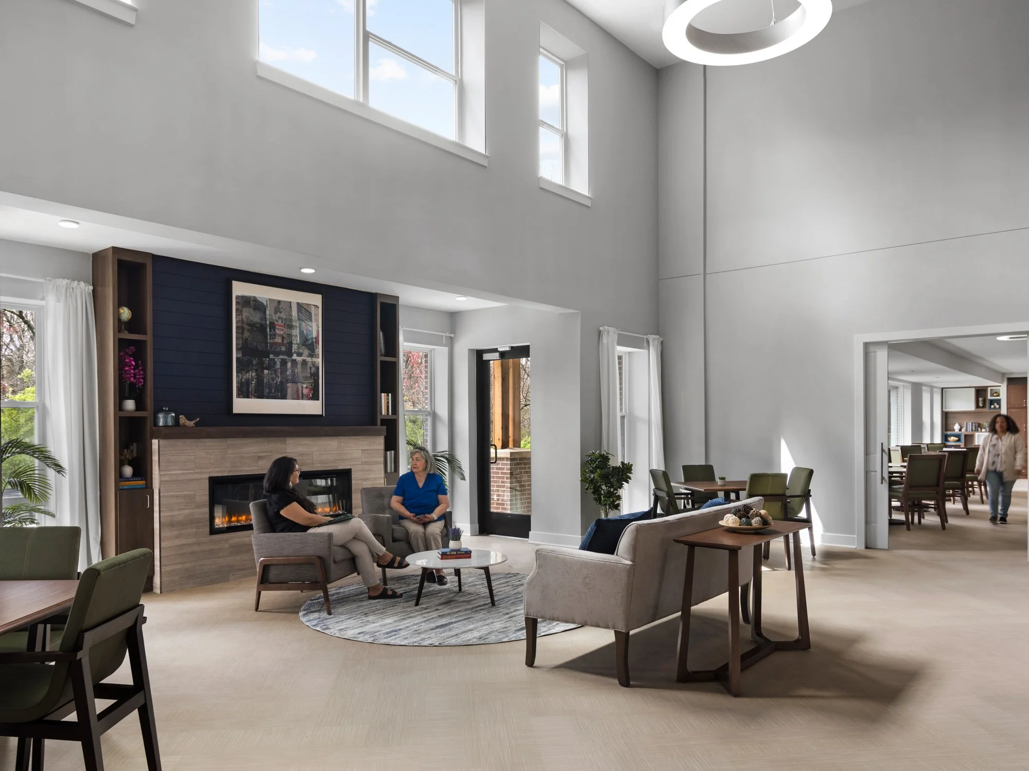 Spacious living room with high ceilings, large windows, white walls, modern fireplace, and minimal decor, with two women sitting and talking, and a woman walking through the dining area in the background.
