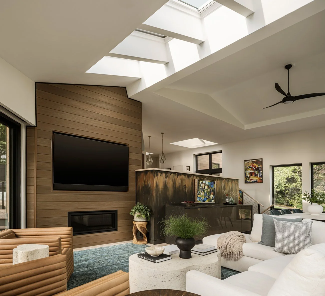 Modern living room with a wood accent wall, large flat-screen TV, skylights, ceiling fan, white sofa with pillows, coffee table with plants and books, artwork on the walls, and large windows showing greenery outside.