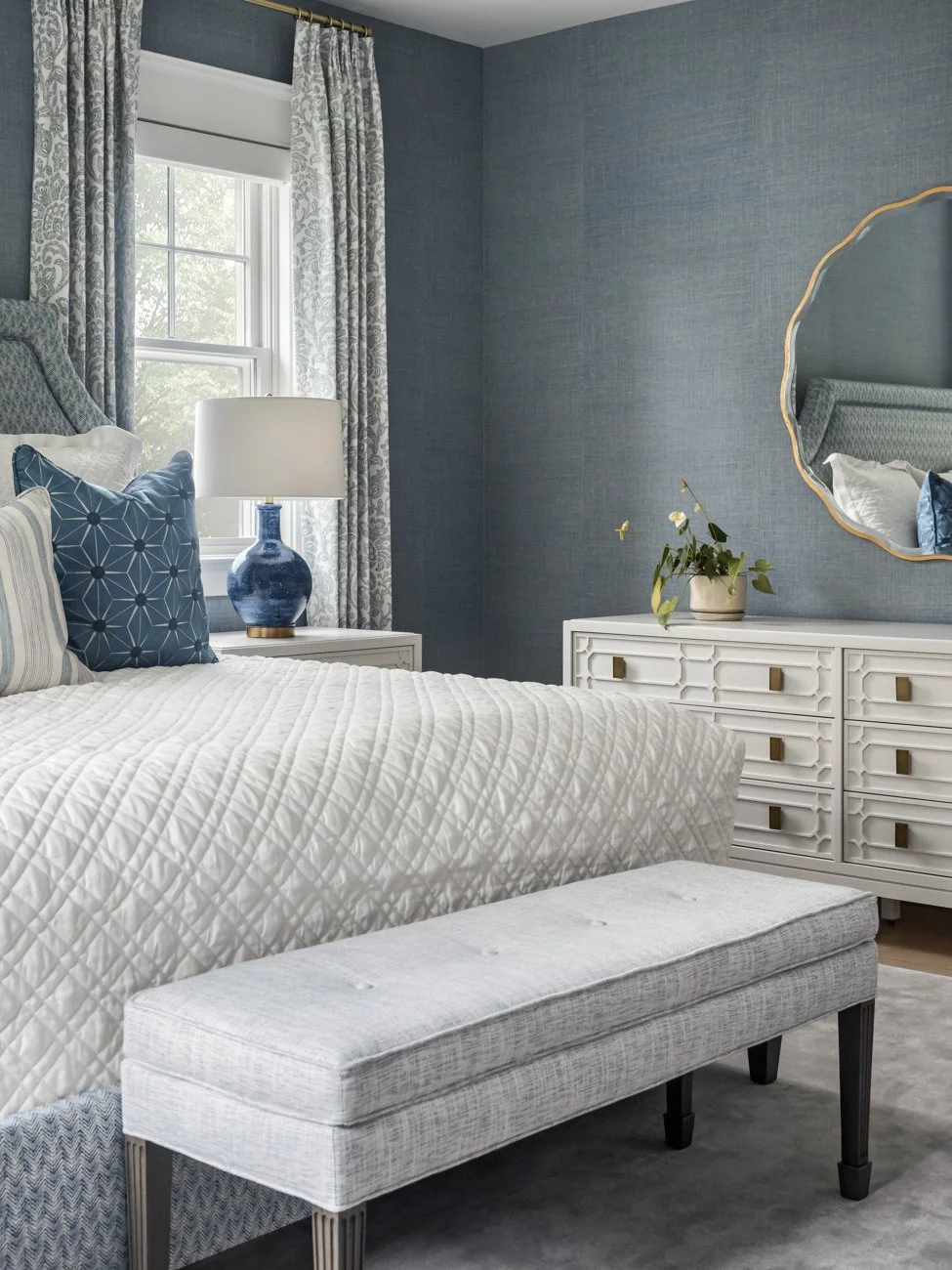 A bedroom with a window, white bed, blue decorative pillows, a white quilt, a gray upholstered bench, a white dresser with a potted plant, a big round mirror, light-colored curtains, and blue textured wallpaper.