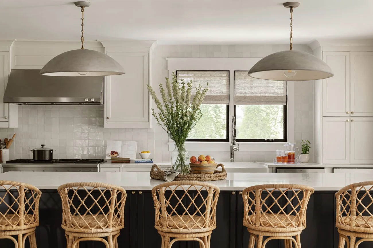 Bright kitchen with white cabinetry, black lower cabinets, a large white countertop island, wicker chairs, and pendant lighting. A large vase with white flowers and a bowl of peaches are on the island, with a window behind the sink showing greenery o