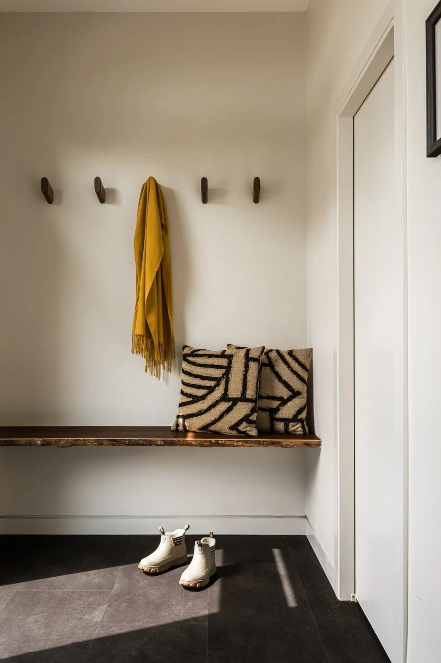 A hallway with a wall featuring four wooden coat hooks, one holding a mustard yellow scarf. Below the hooks, a wooden bench with two decorative pillows with black and beige geometric patterns. On the black tiled floor, a pair of white rain boots. To 