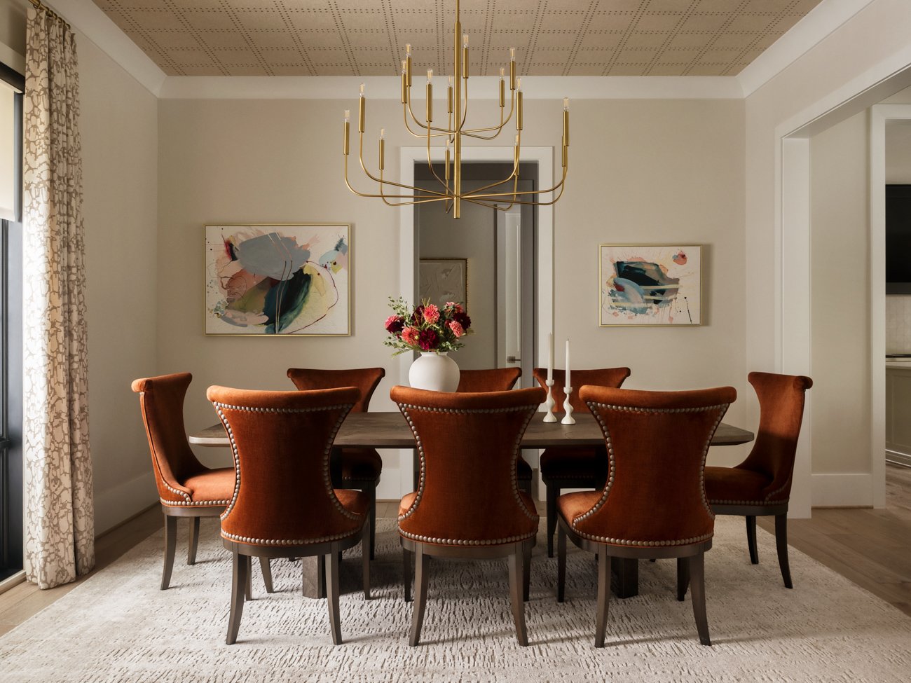Dining room with a dark wooden table, eight brown leather chairs, a white vase with pink flowers, white candlesticks, abstract art on the walls, a gold chandelier, beige curtains, and a light-colored area rug.