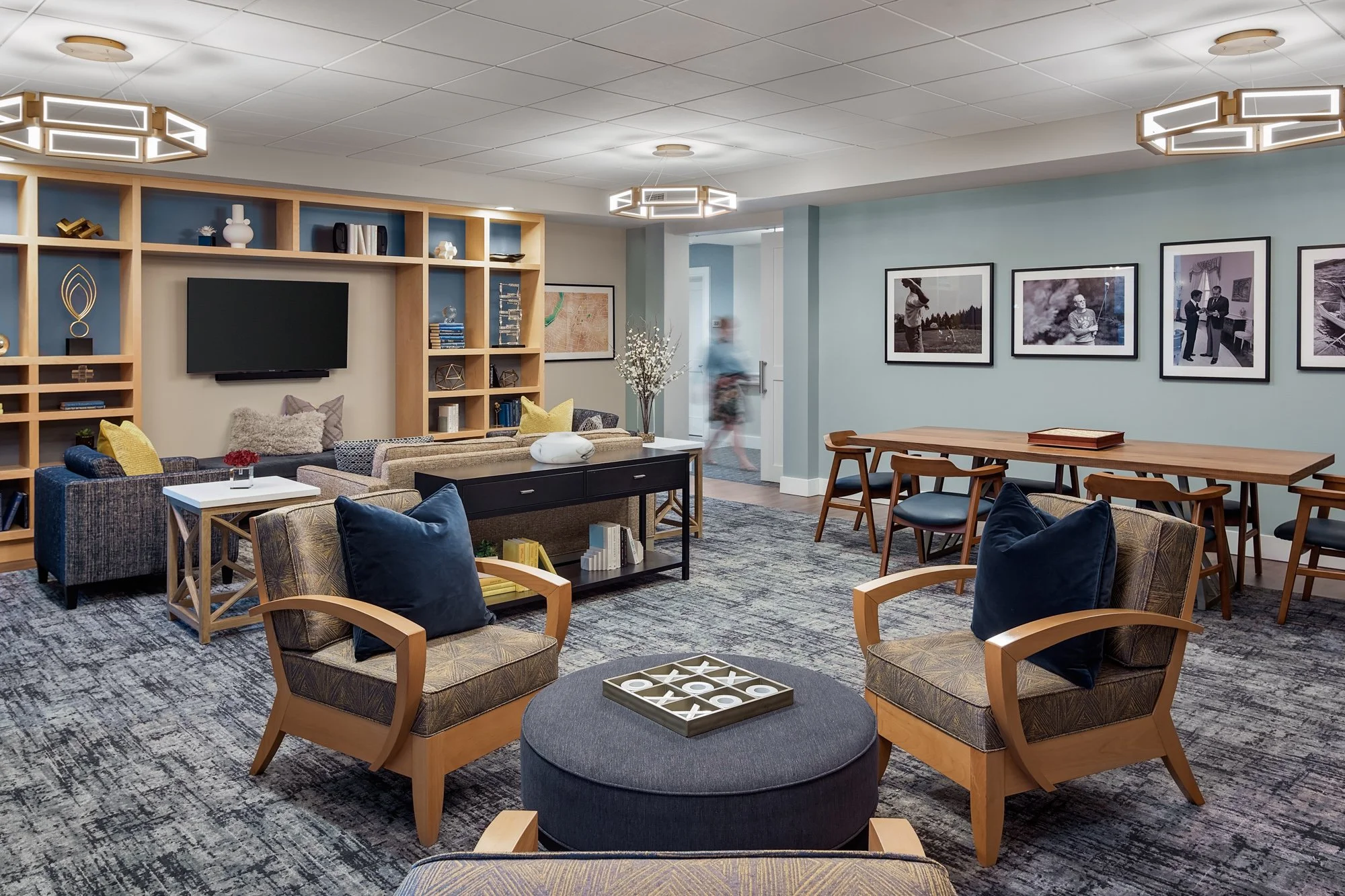 Modern lounge with multiple chairs, a tv, wall art, bookshelves, and a person walking in the background.