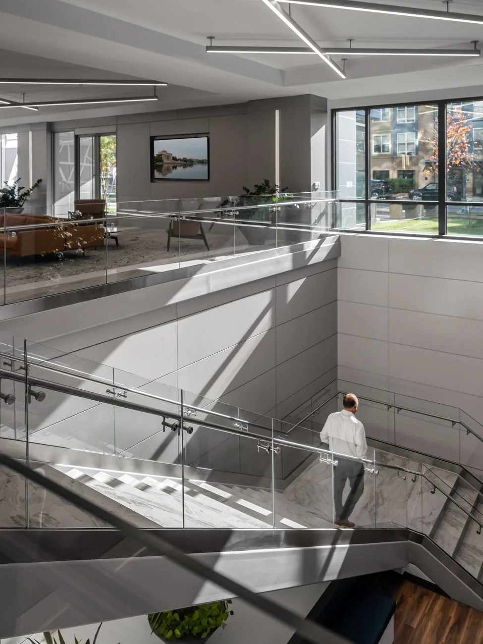 A modern multi-level office interior with glass railings, a staircase, large windows, and minimalistic furniture. A man in business attire is walking down the stairs.