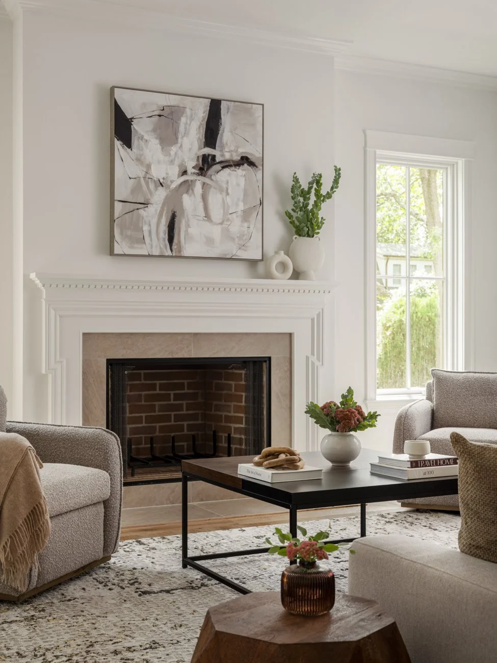 Living room with a white fireplace, abstract black and white artwork above it, and a black coffee table with books and a vase of flowers. There are beige sofas, a window with trees outside, and decorative vessels on the mantle.