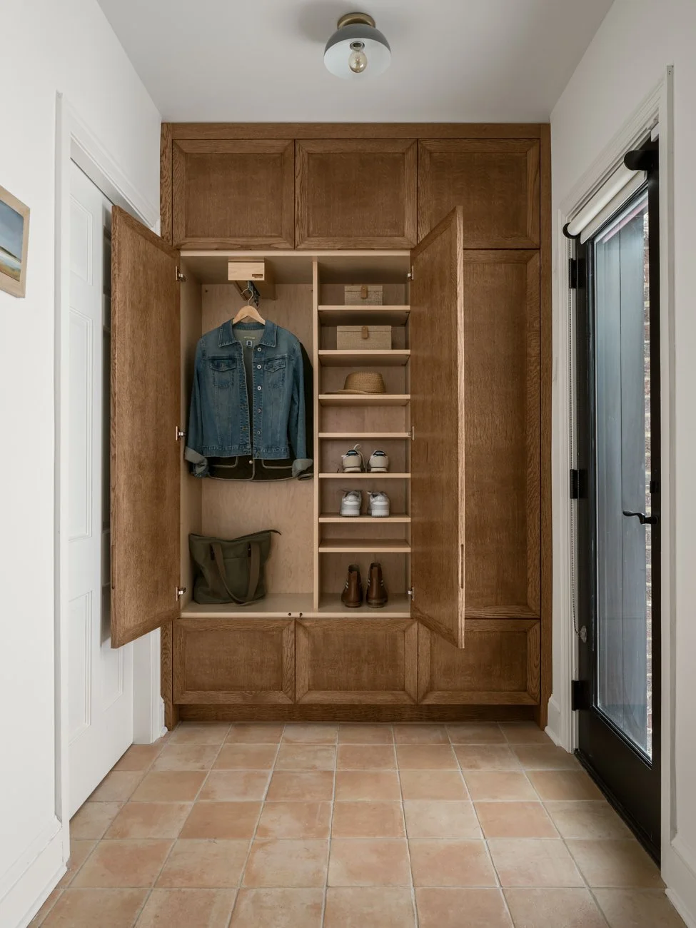 Open wooden closet with hanging denim jacket and a bag, shoe compartments with sneakers and boots, a hat on shelf, and a door to the outside.