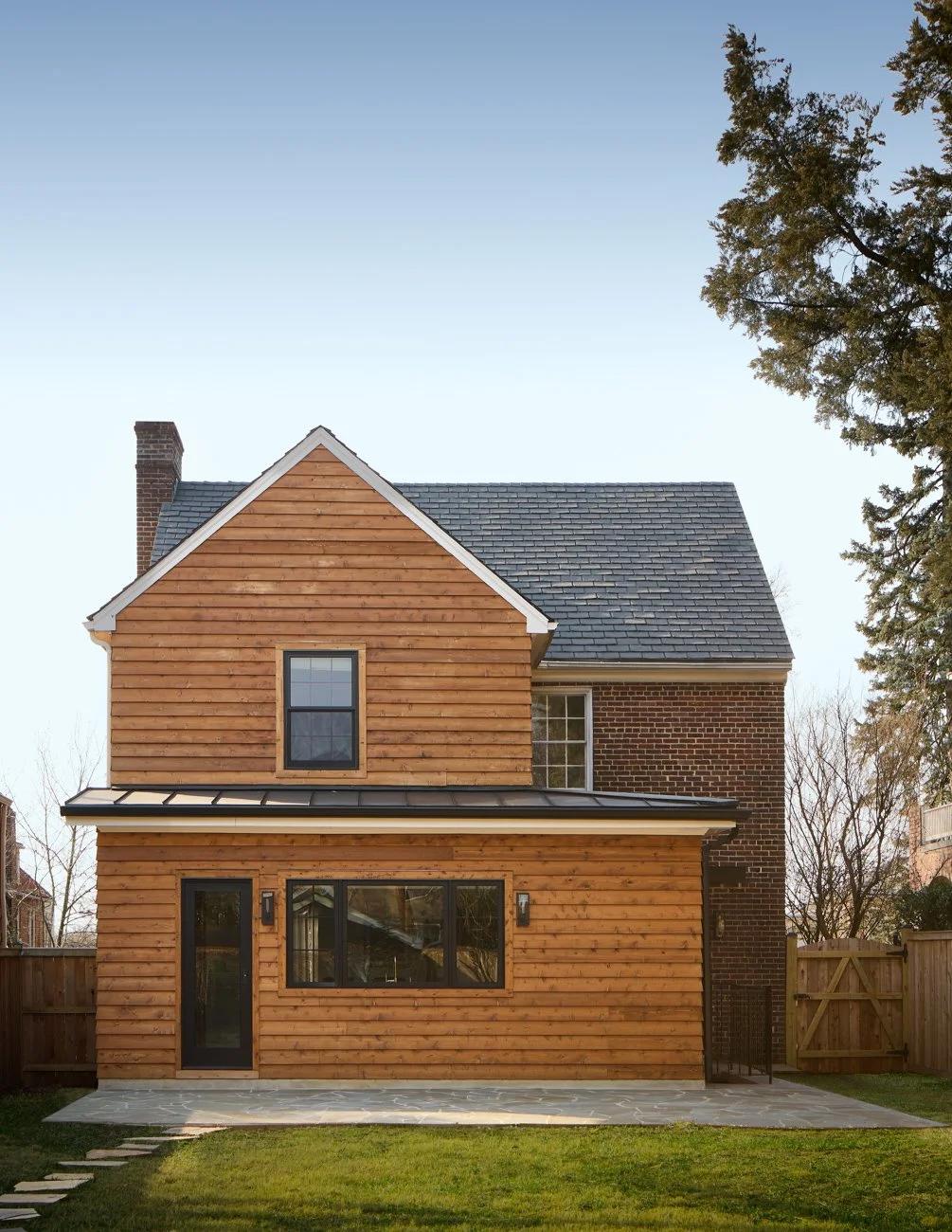 Rear view of a modern two-story house with wood siding on the first floor and bricks on the second, a sloped roof, and a small patio. A wooden fence surrounds the yard, and trees are visible in the background.