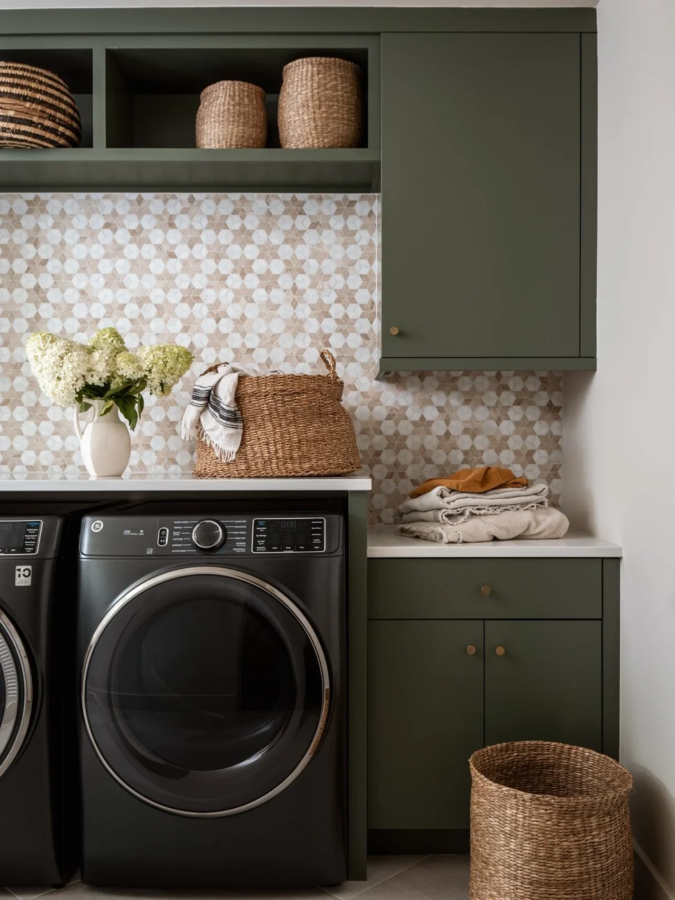 Laundry room with green cabinets, a washing machine, a white counter with folded clothes, a basket, a vase of white flowers, and woven baskets on shelves and the floor.