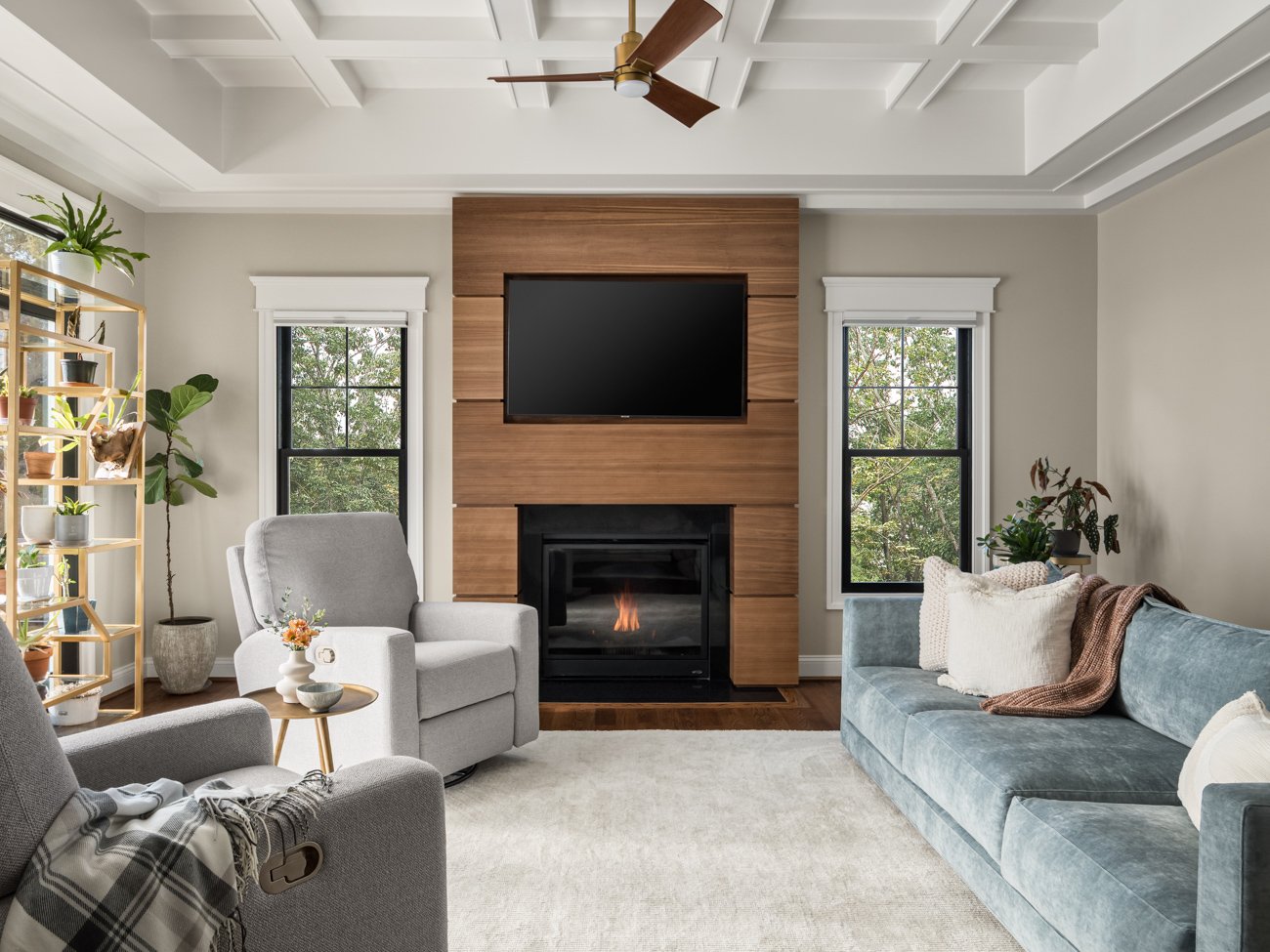 Living room with light gray and blue furniture, a fireplace, a flat-screen TV, windows with trees outside, plants, and a ceiling fan