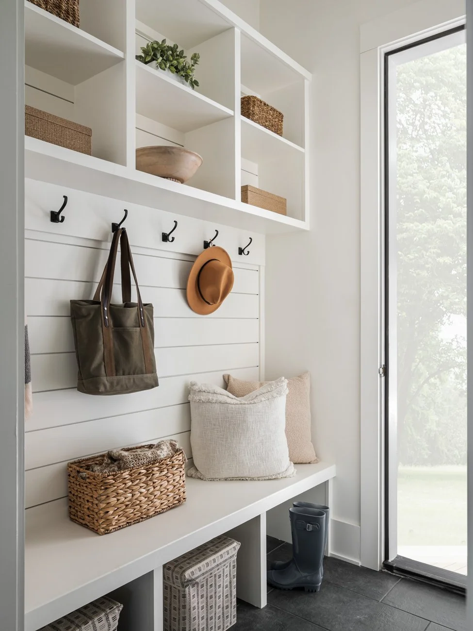 Entryway with white wooden bench, hanging hooks with a bag and a hat, shelves with storage baskets, pillows, and a pair of rain boots near a glass door with natural light.