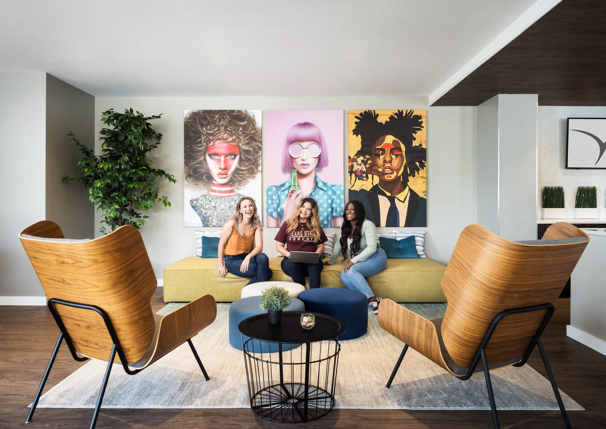 Three women sitting on a yellow sofa in a modern living room, laughing and talking. Behind them are three colorful art portraits on the wall, a large plant to the left, and two wooden chairs in the foreground facing the sofa. A black round coffee tab