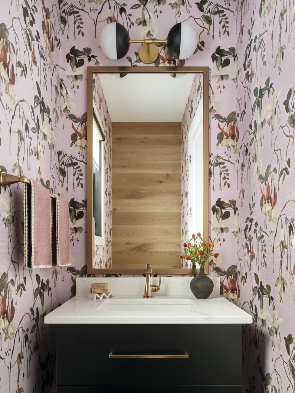 A bathroom vanity with a white countertop, black cabinet, and gold handle, featuring a brass faucet, a black vase with orange flowers, and a mirror. The pink floral wallpaper has dark green, cream, and reddish accents. A wall-mounted towel rack holds