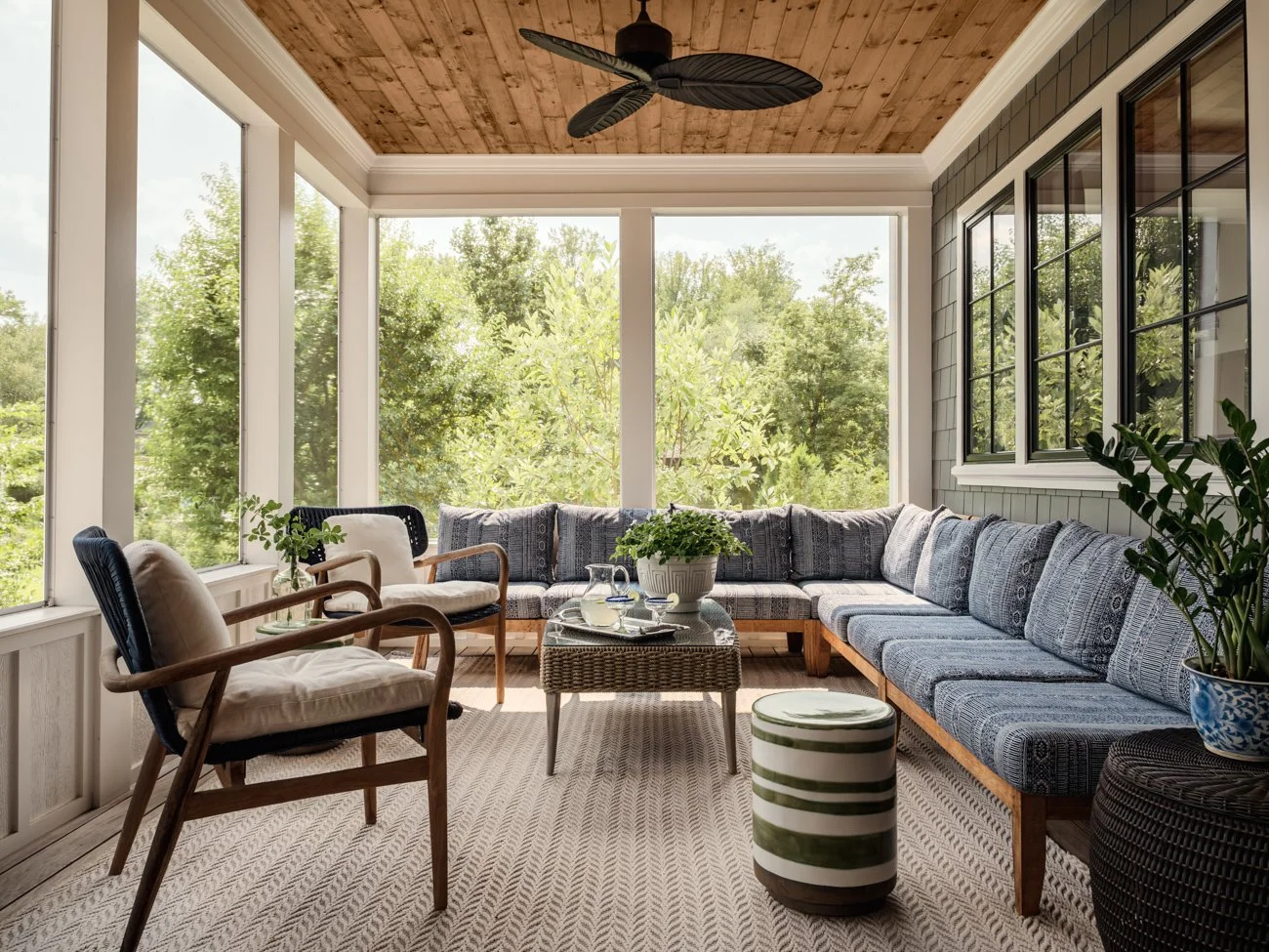 Sunroom with large glass windows, wooden ceiling with a ceiling fan, and a mix of cushioned chairs and a long sectional sofa with patterned blue cushions. There are plants on tables and the floor, a wicker table with a tray, and an area rug.
