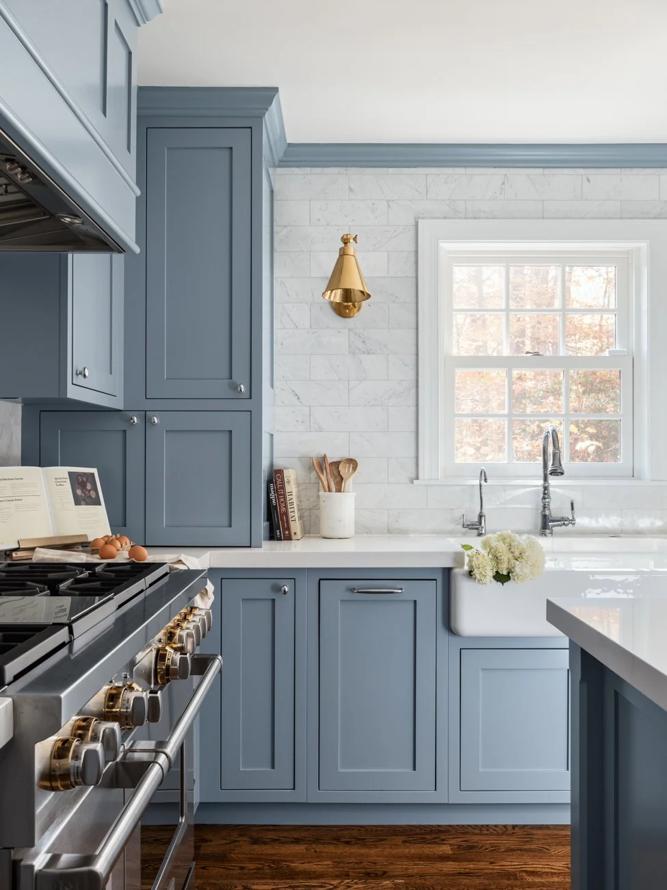 A modern kitchen with blue cabinets, a white marble countertop, and a farmhouse sink. There is a window above the sink with a view of trees, and a gold wall sconce on the tiled white marble wall. Cooking utensils and books are on the countertop.