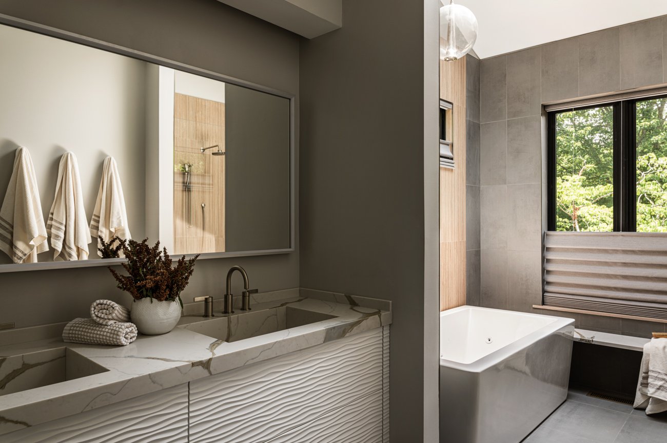 Modern bathroom with a white marble vanity, beige towels, a mirror, a walk-in shower, and a large bathtub next to a window showing greenery outside.