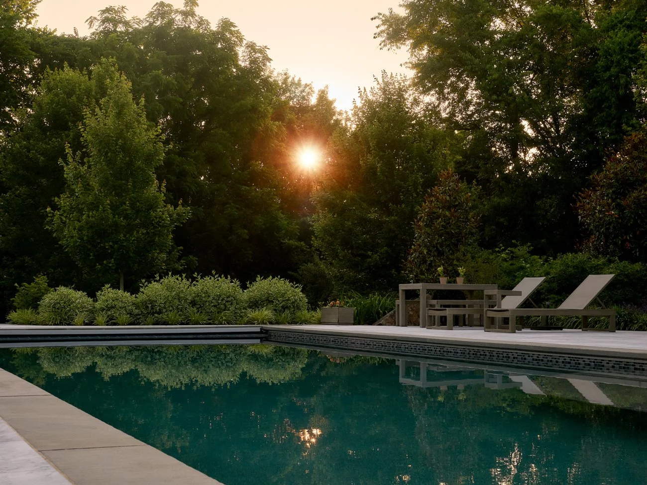 An outdoor swimming pool with lounge chairs and a small table on a deck, surrounded by lush green trees and plants at sunset.