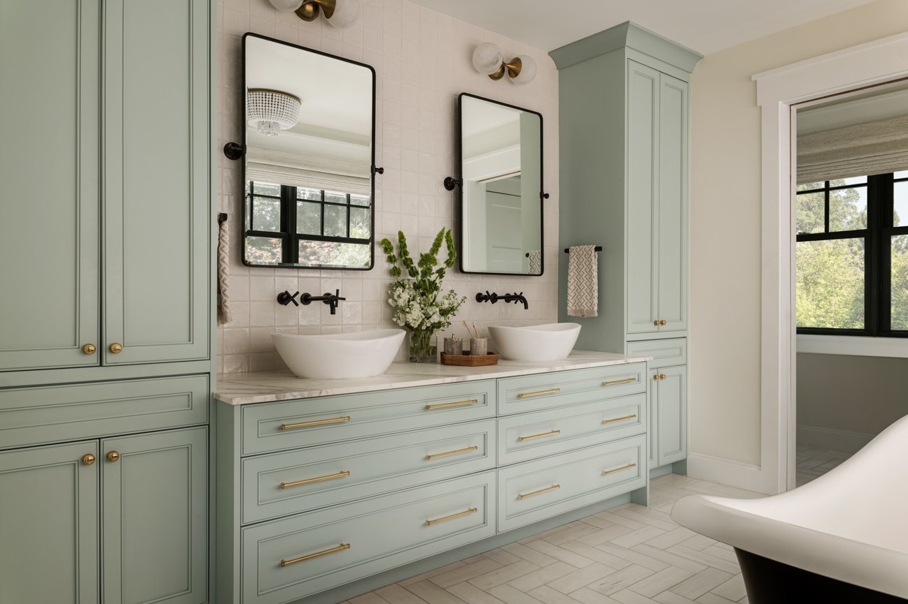 A modern bathroom with a double vanity with light green cabinets, two white vessel sinks, black wall-mounted faucets, two rectangular mirrors, and a bouquet of white flowers on the countertop. There is a window reflecting natural light and a separate