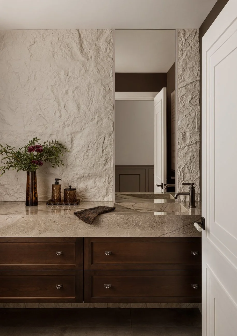 A bathroom vanity with a beige marble countertop, dark wooden drawers, a large mirror, and decorative items including a vase with purple flowers, soap dispensers, and a small tray. There's a textured stone wall behind the vanity and a door to the rig