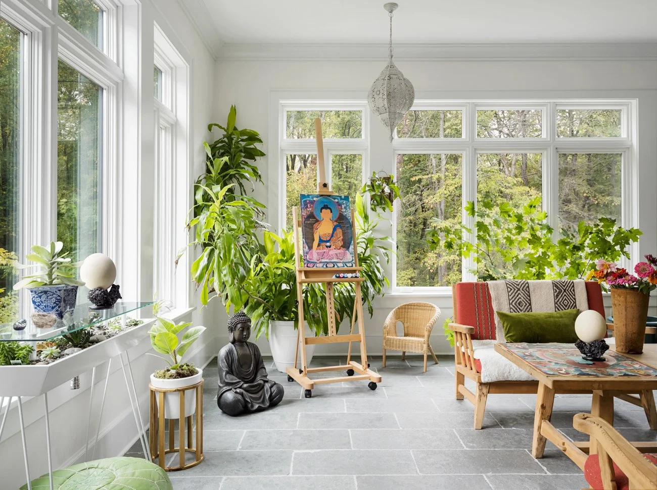 Bright sunlit living room with large windows, houseplants, a Buddha statue, and a wooden easel with a colorful painting of a woman in traditional attire.