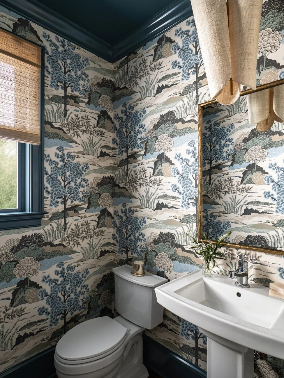 A small bathroom with wallpaper featuring a nature scene in blue, gray, and beige tones, a white toilet, a white sink with a faucet, a rectangular mirror with a gold frame, a vase with greenery on the sink, a small candle on top of the toilet, a wind