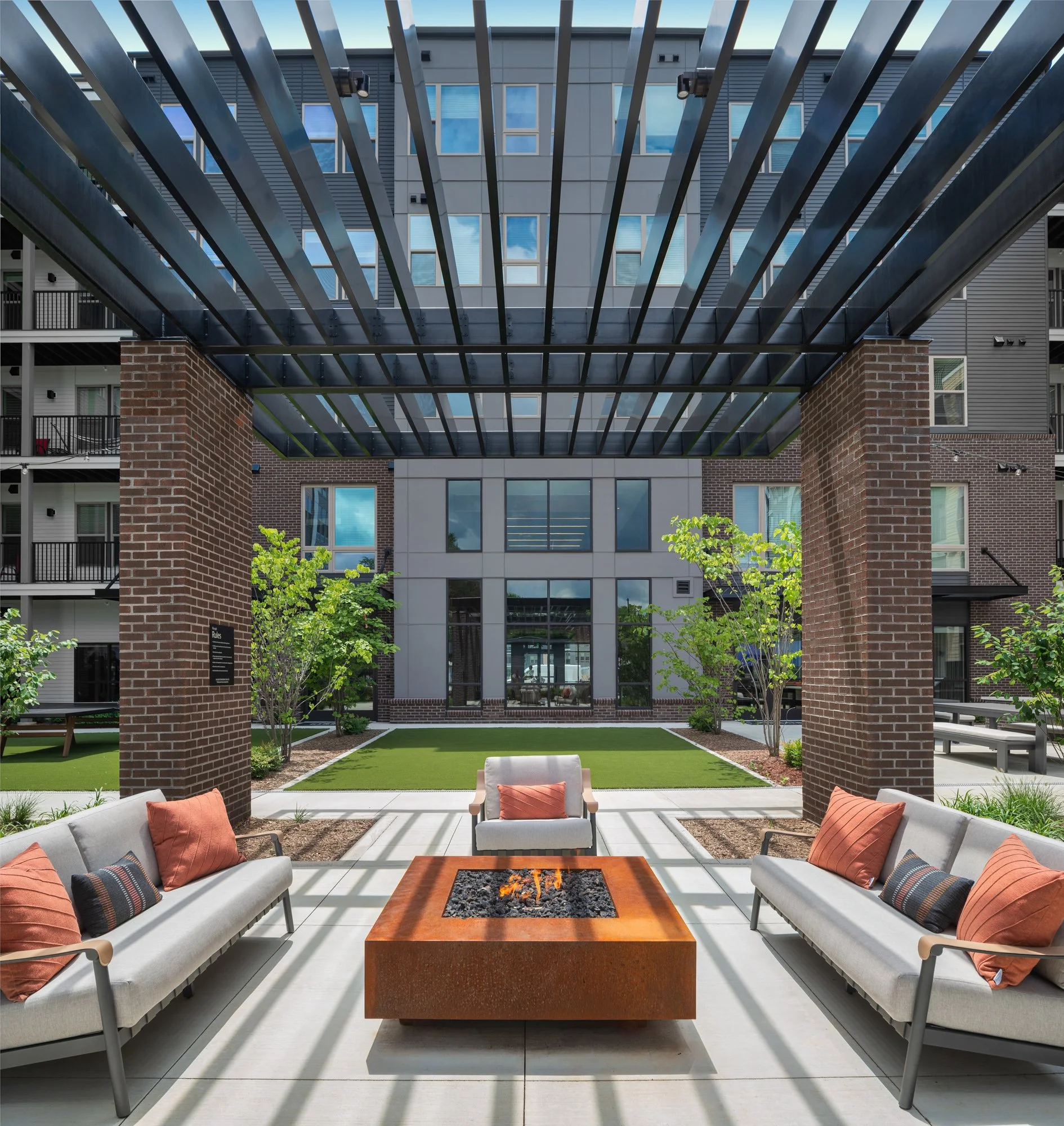 Outdoor patio with modern seating area featuring three sofas with cushions around a fire pit, under a metal pergola, with a landscaped courtyard and apartment building in the background.