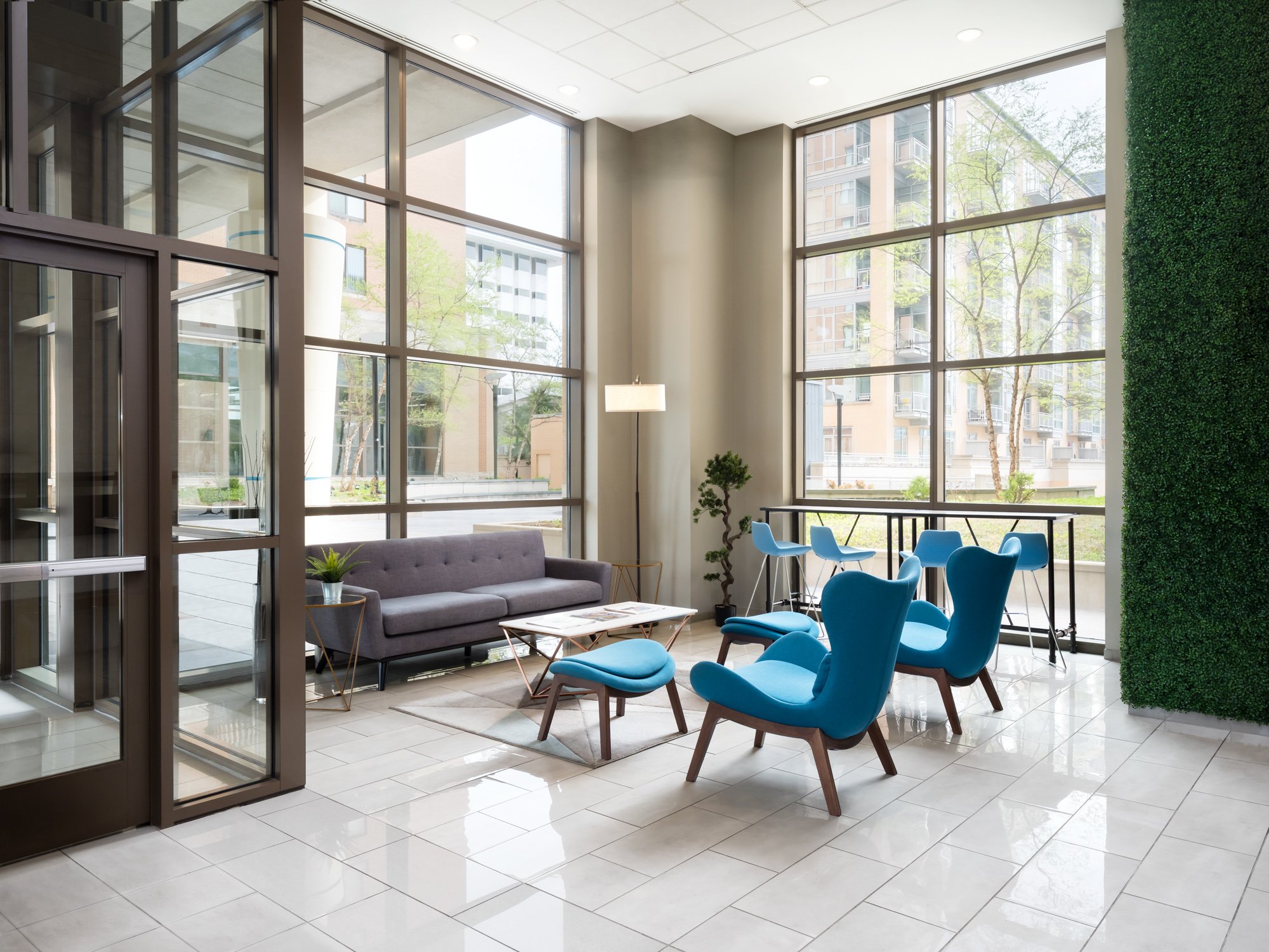 Modern lounge area with large floor-to-ceiling windows, blue chairs, a purple sofa, a small coffee table, a tall green plant, and a wall with green foliage decor.