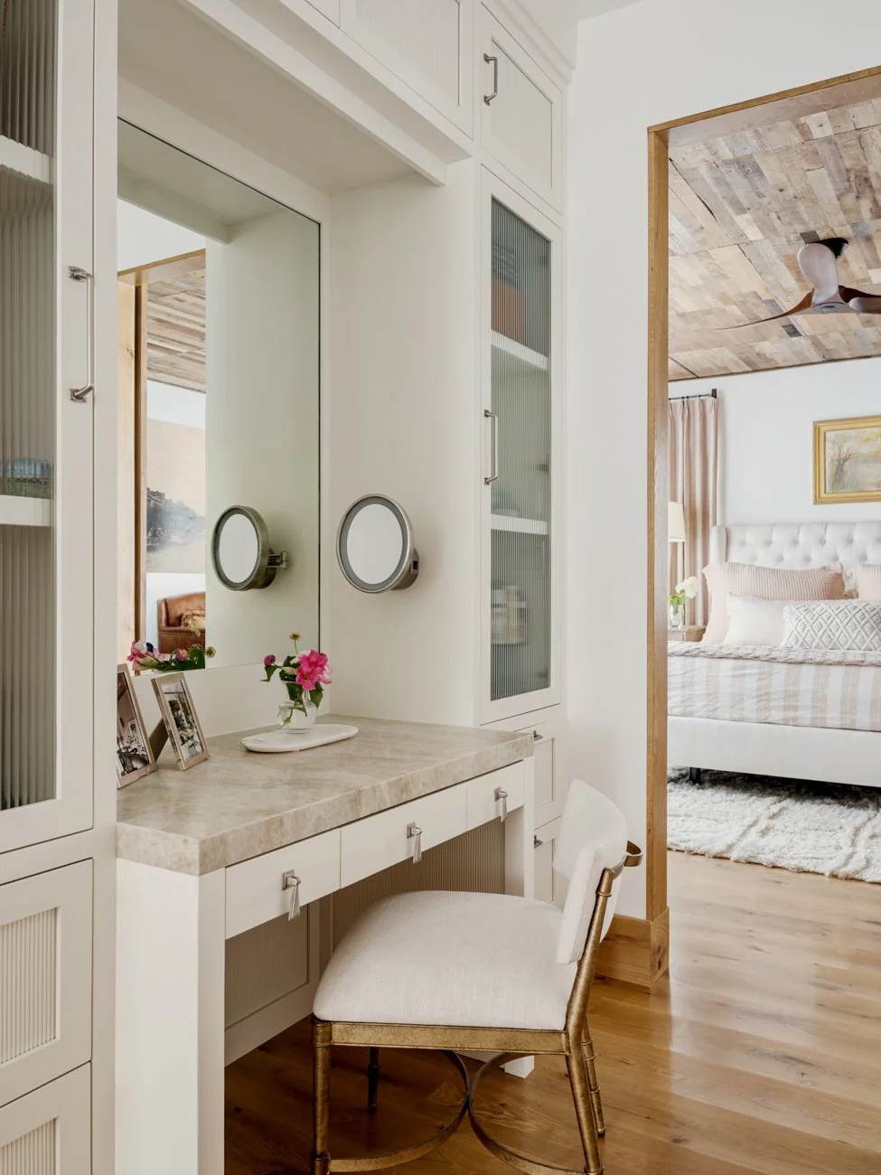 A bedroom with a vanity desk, mirror, framed photos, pink flowers, and a white upholstered chair in front of the mirror, with a bedroom visible through an open doorway.