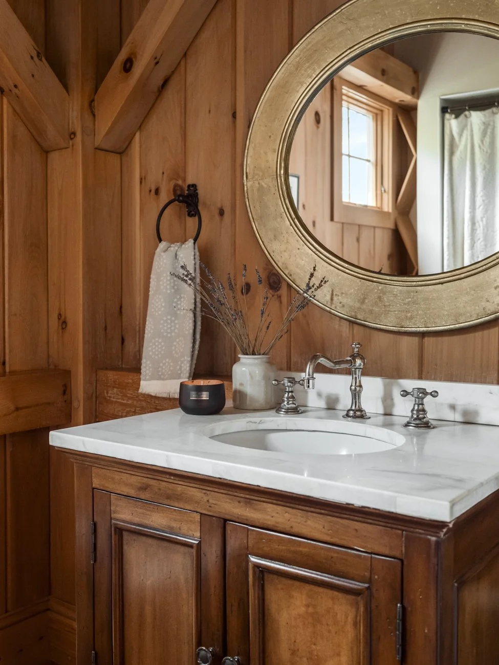 Wood-paneled bathroom vanity with a white marble countertop, oval mirror, and decor including a vase with dried lavender and a candle.