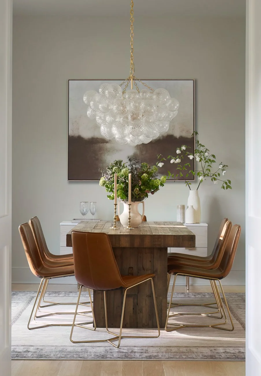 A dining room with a rustic wooden table, six tan leather chairs with gold legs, a modern chandelier made of clear glass globes hanging above, a white sideboard with decorative vases and tall candlesticks, a large abstract painting on the wall, and a